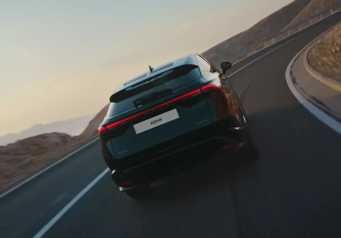 Vista traseira do Nissan Ariya em movimento numa estrada de montanha, destacando a barra de luzes LED contínua e o design coupé do SUV elétrico