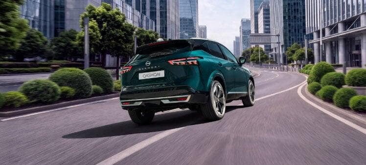 Vista traseira de um Nissan Qashqai SUV de cor verde esmeralda, a circular numa estrada citadina ladeada por vegetação e edifícios contemporâneos.