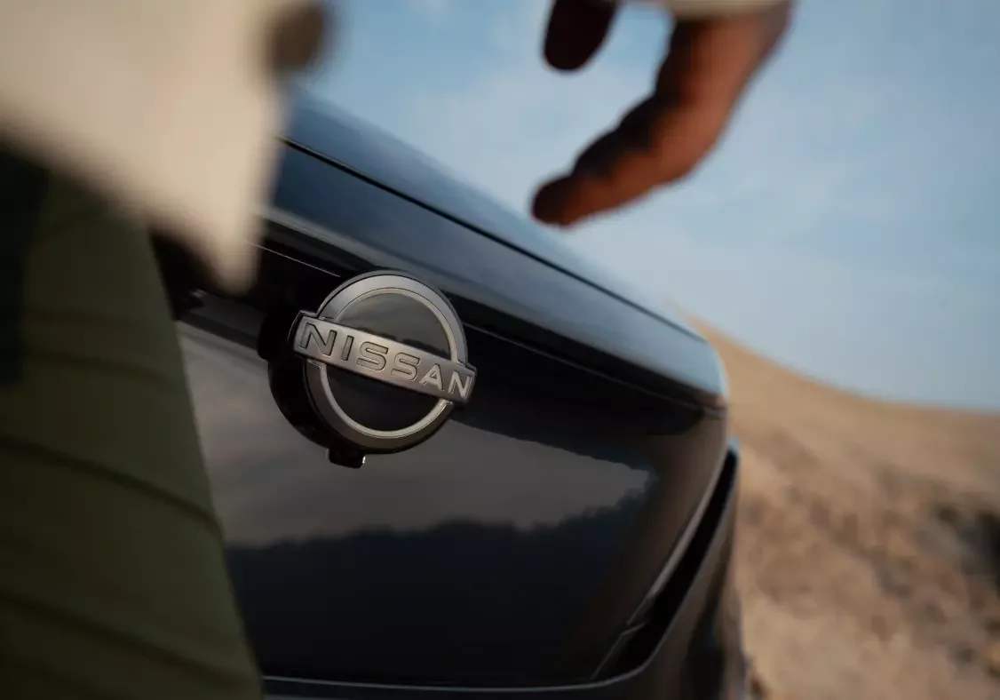 Pormenor do emblema iluminado da Nissan na frente do Ariya, com mão a aproximar-se em ambiente de paisagem desértica