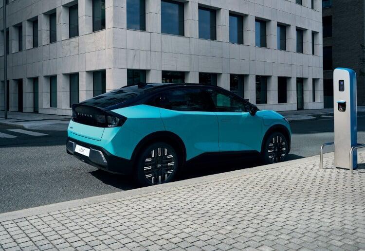 Vista lateral do novo Nissan LEAF, em azul teal. O carro encontra-se parado numa estrada urbana, com um edifício cinzento em plano de fundo. 