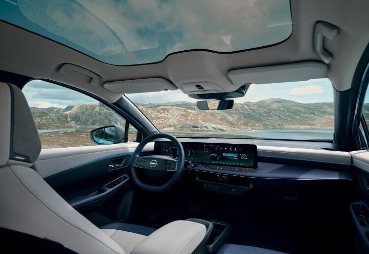 Vista interior do novo Nissan LEAF, com detalhe para os dois ecrãs de 14,3’’, acabamentos de estofo brancos e ainda o teto panorâmico com funções de escurecimento. No exterior vemos montanhas, céu azul e um espelho de água. 