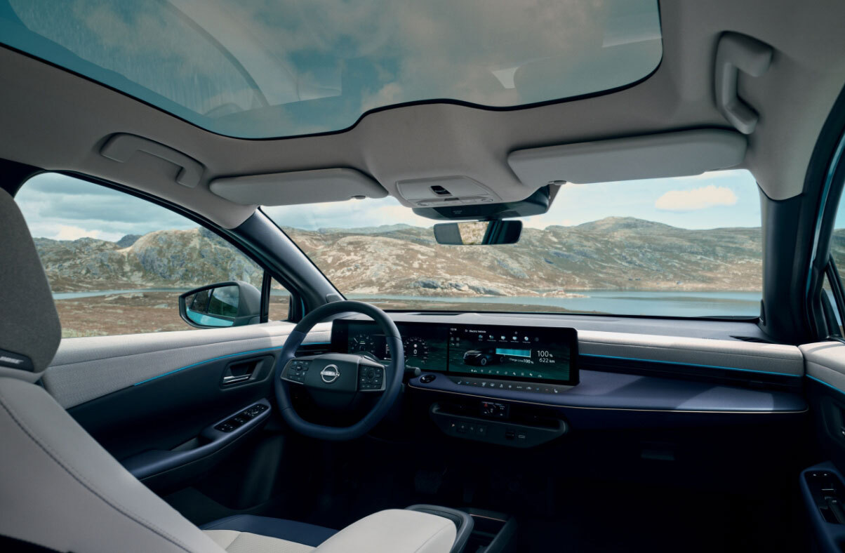 Vista interior do novo Nissan LEAF, com detalhe para os dois ecrãs de 14,3’’, acabamentos de estofo brancos e ainda o teto panorâmico com funções de escurecimento. No exterior vemos montanhas, céu azul e um espelho de água.