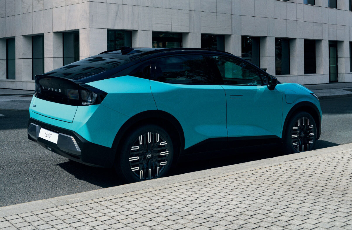 Vista lateral do novo Nissan LEAF, em azul teal. O carro encontra-se parado numa estrada urbana, com um edifício cinzento em plano de fundo. 