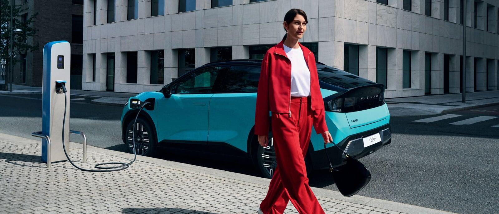 - Mulher jovem vestida com um conjunto vermelho caminha junto ao novo LEAF. O carro está a carregar, num ponto de carregamento citadino, com um edifício cinzento em plano de fundo. 