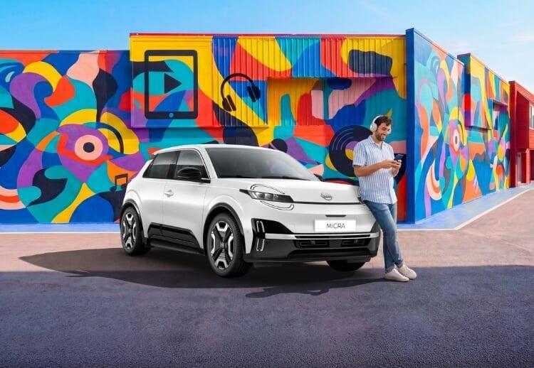 Nissan Micra elétrico branco estacionado diante de mural colorido com ícones de mídia e fones de ouvido; homem com fone de ouvido mexendo no celular ao lado do carro.