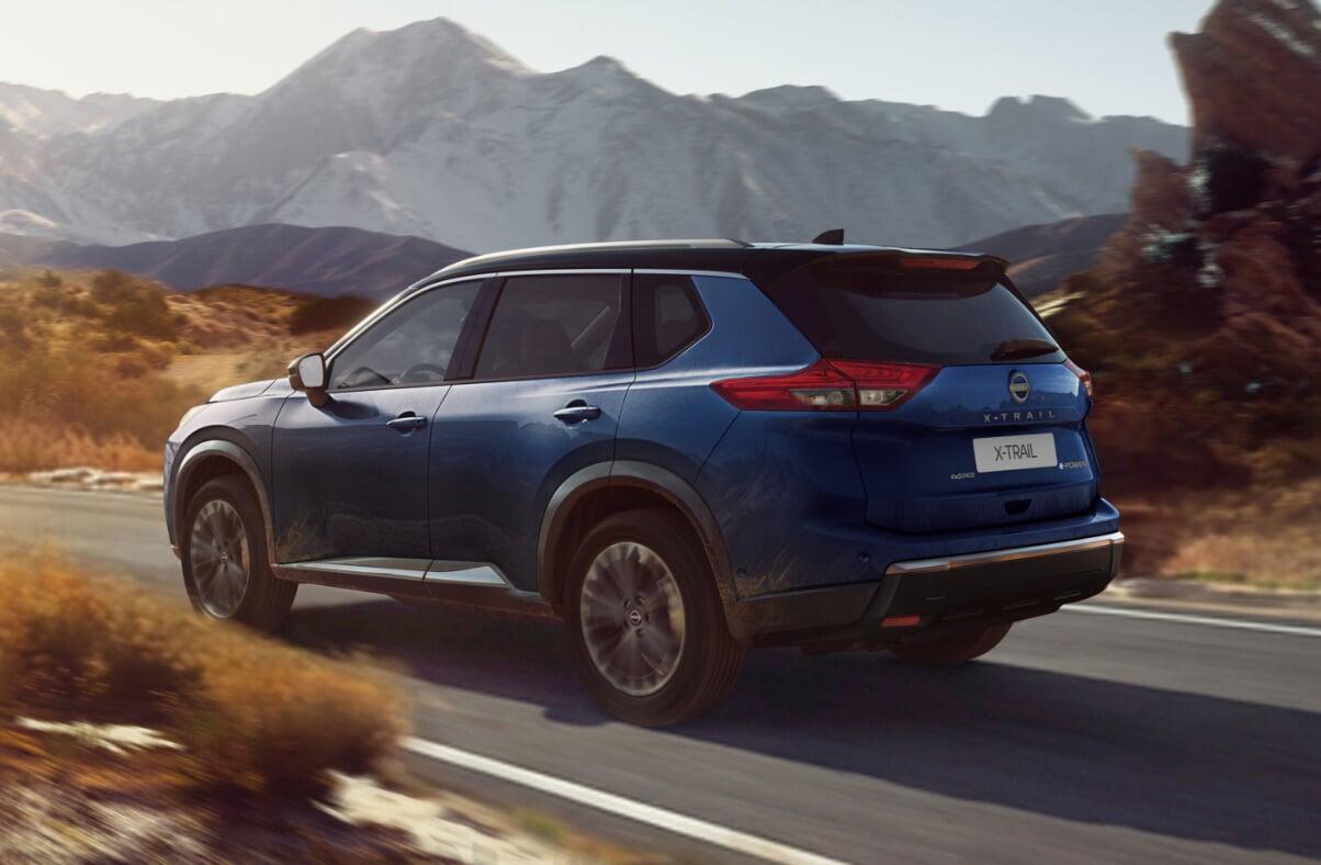 Nissan X-Trail azul visto de trás em movimento numa estrada, com montanhas rochosas ao fundo