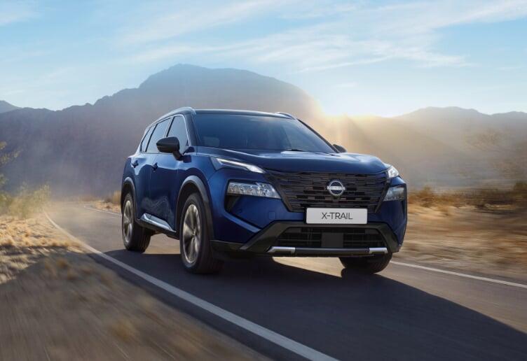 Nissan X-Trail azul visto de frente em movimento numa estrada de montanha, com o sol a nascer ao fundo
