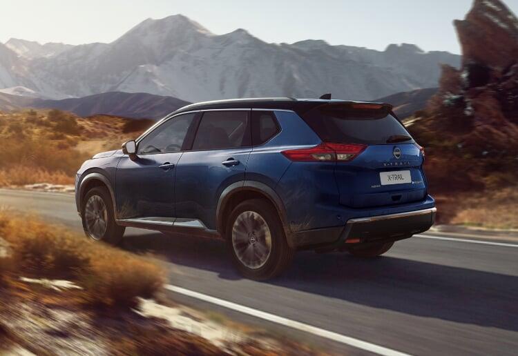Nissan X-Trail azul visto de trás em movimento numa estrada, com montanhas rochosas ao fundo