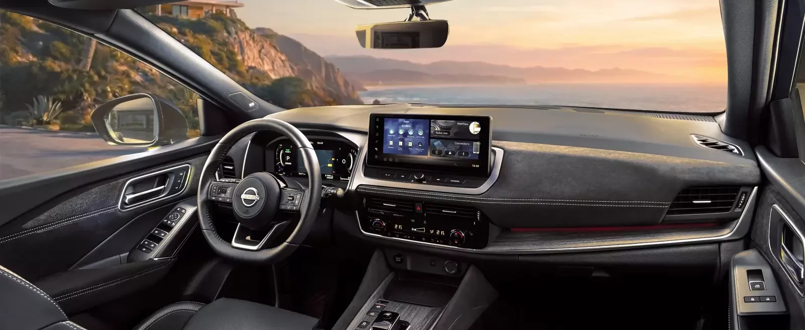 Nissan Qashqai e-POWER interior com volante multifunções, painel digital e ecrã central de infotainment, com vista para uma estrada costeira ao pôr do sol.